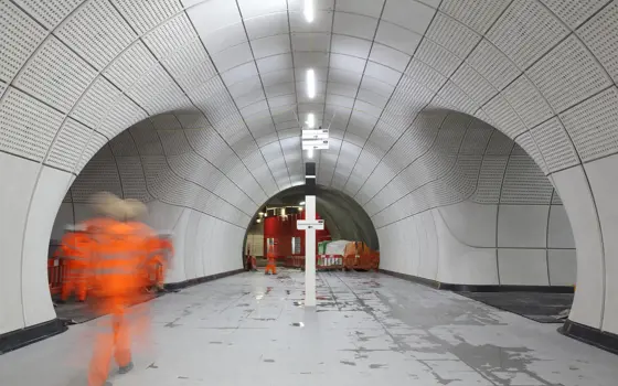 The inside of the underground Elizabeth Line station while it was under construction with glass fibre reinforced concrete cladding panels lining the walls. A blurry worker in construction gear is walking through the tunnel. 