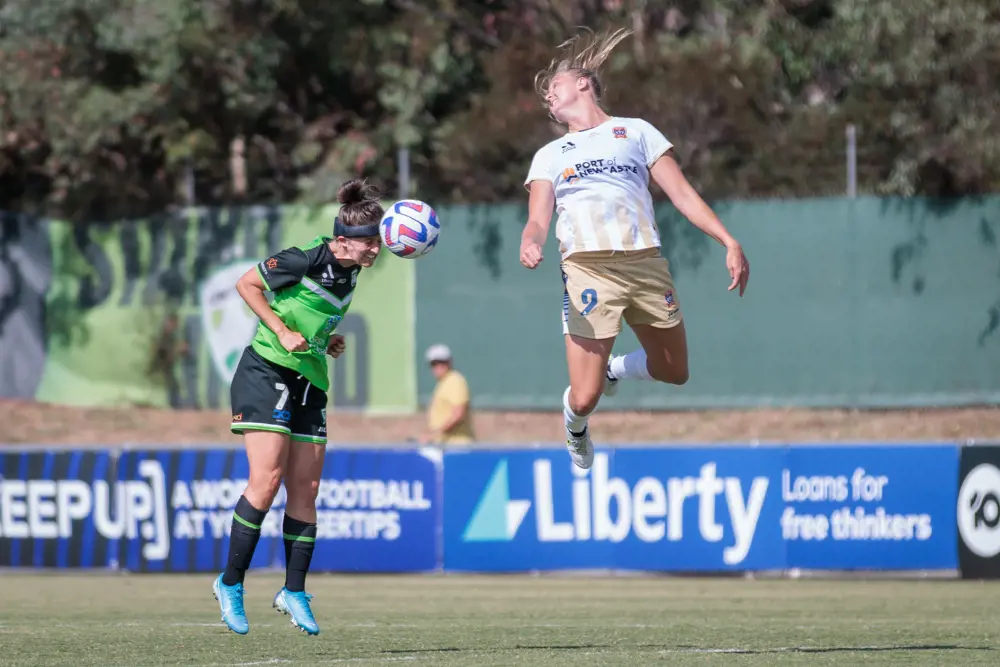 A white woman footballer with brown hair tied in a ponytail heads a ball on an outdoor sports pitch. She is wearing a green and black football kit with blue football boots, and also wears a black protective sports headband. A second white woman footballer with blonde hair tied in a ponytail is slightly ahead of her and mid-jump, having just missed the football. She wears a cream and white football kit,