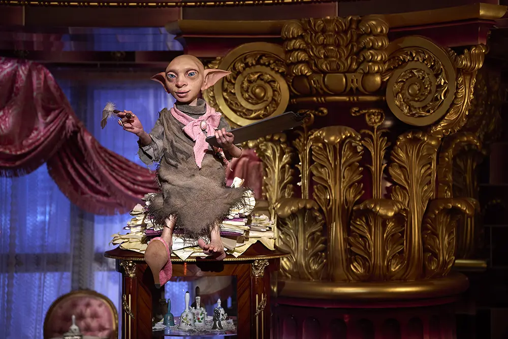 An animatronic model of an elf-like creature wearing a sack sits on a pile of papers atop a display cabinet in front of a gold ornate column