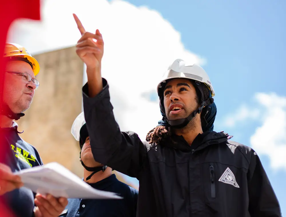 A person wearing a safety helmet and outdoor gear gestures upwards while standing with other helmeted team members holding papers at an outdoor work site.