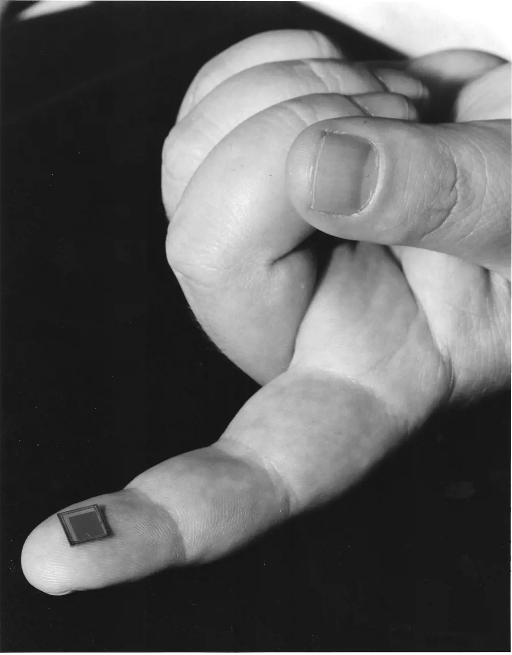 A small square camera sensor resting on someone's fingertip in greyscale.