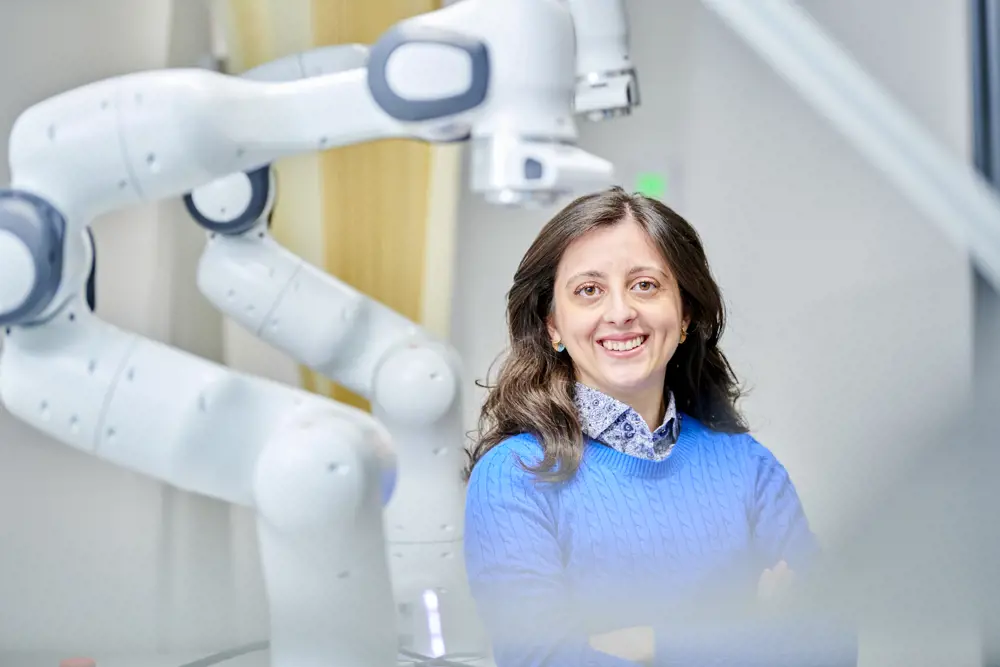 A woman with brown hair and brown eyes smiling at the camera, wearing a blue jumper and standing in front of large robotic arms.