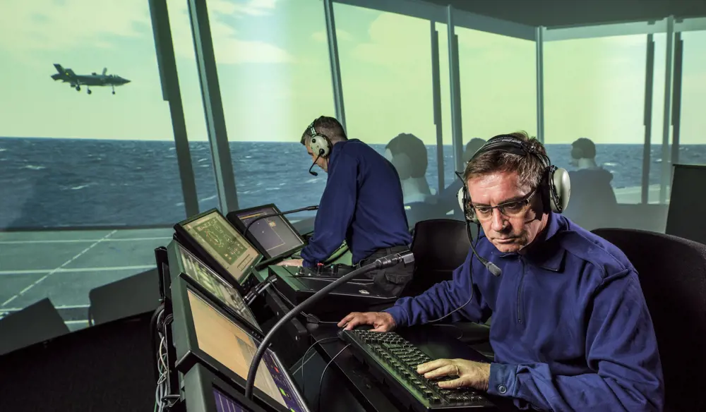 A virtual realistic image of the ship's flying control tower on the simulator, with two males in headsets looking at screens and a fighter plane can be seen outside the window.