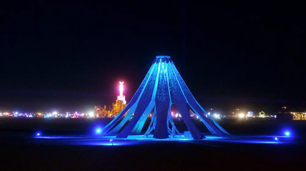 A tent-like pavilion structure made of glowing blue petals with openings in between, shown at night, with other glowing structures seen in the distance.