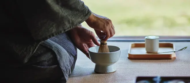 Hands whisking tea in a ceramic bowl during a traditional Japanese tea ceremony, with utensils on a tatami mat.