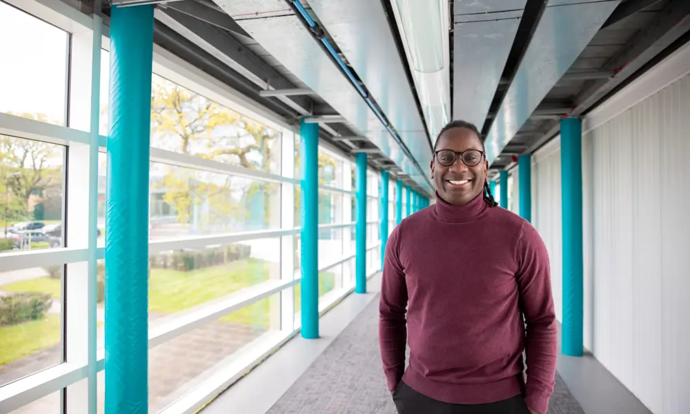 James Dornor wearing glasses and a burgundy jumper stands smiling in a bright corridor with turquoise columns and large windows.