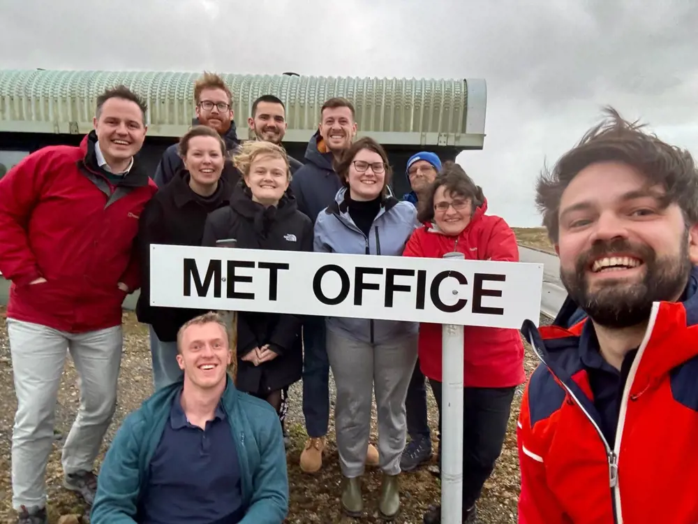 A group of people on a blustery day gathering for a photo around a "Met Office" sign.