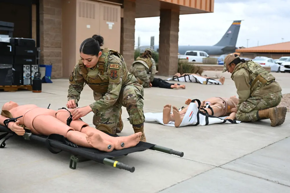 Two women army soldiers kneel on the floor outside, practicing tying a black tourniquet around medical dummies.