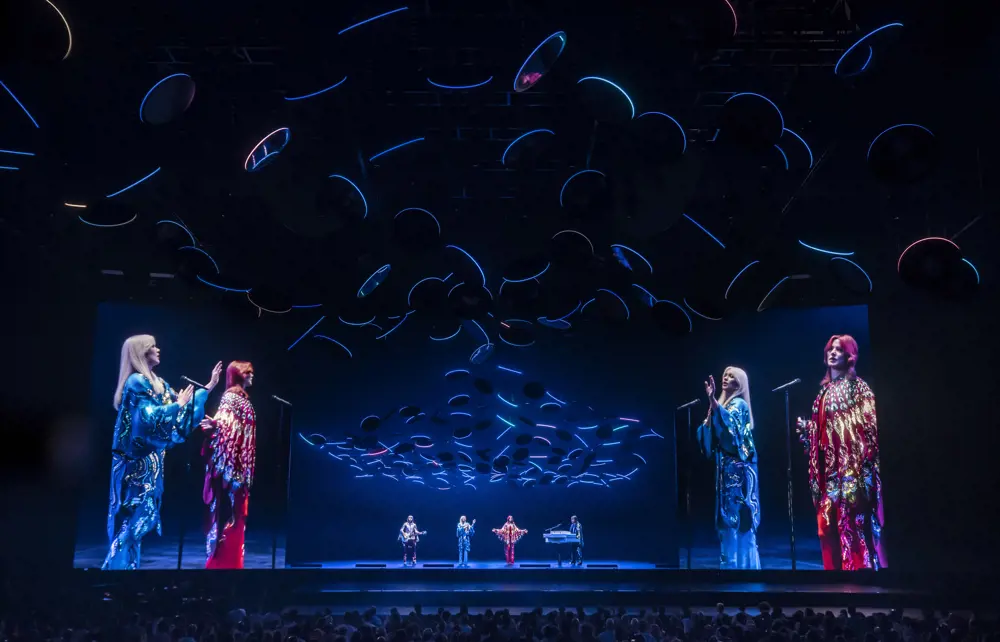 Two singers, a guitarist and a keyboard player wearing elaborately pattered red and blue outfits stand on a stage and perform to a crowd, while subtly illuminated by circular LED lights suspended from the roof