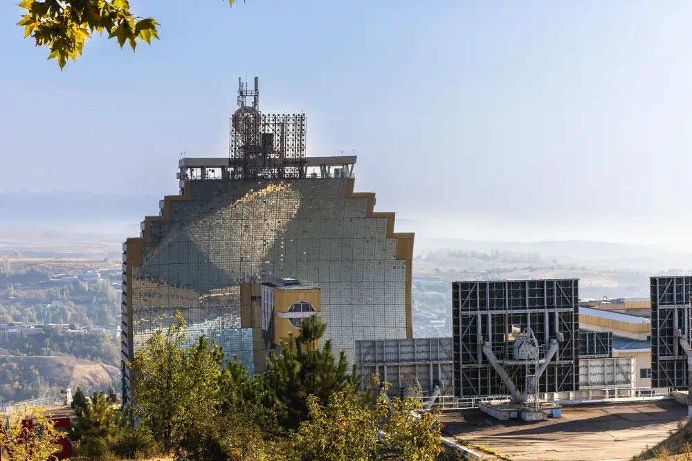 A vast, concave building covered with mirrors glinting in the sun. It is situated on top of the hill with a hazy background of trees, buildings and a blue sky above.