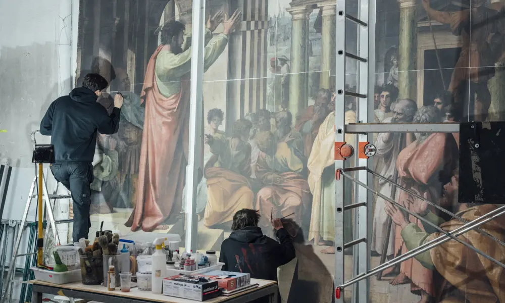 Two artists, one on a ladder and one crouching, who are working on a replica of a Raphael painting next to a table of art supplies.