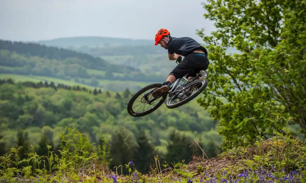 A person with a helmet riding a mountain bike in the air, with rolling green hills in the background.