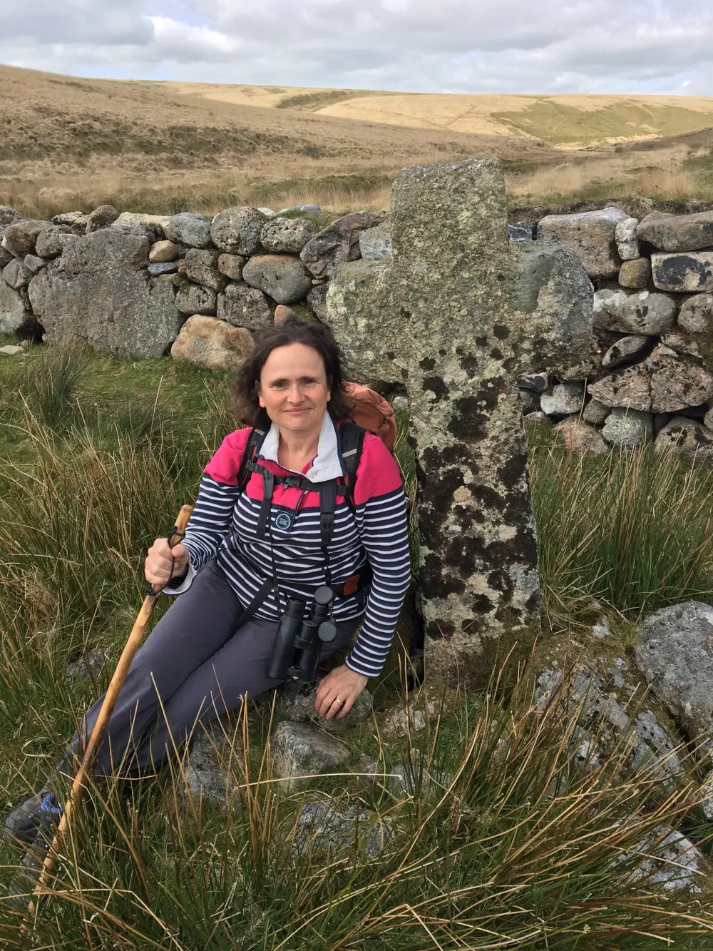 Penny wearing hiking gear with binoculars around her neck, sitting on a rock with moorland in the background.