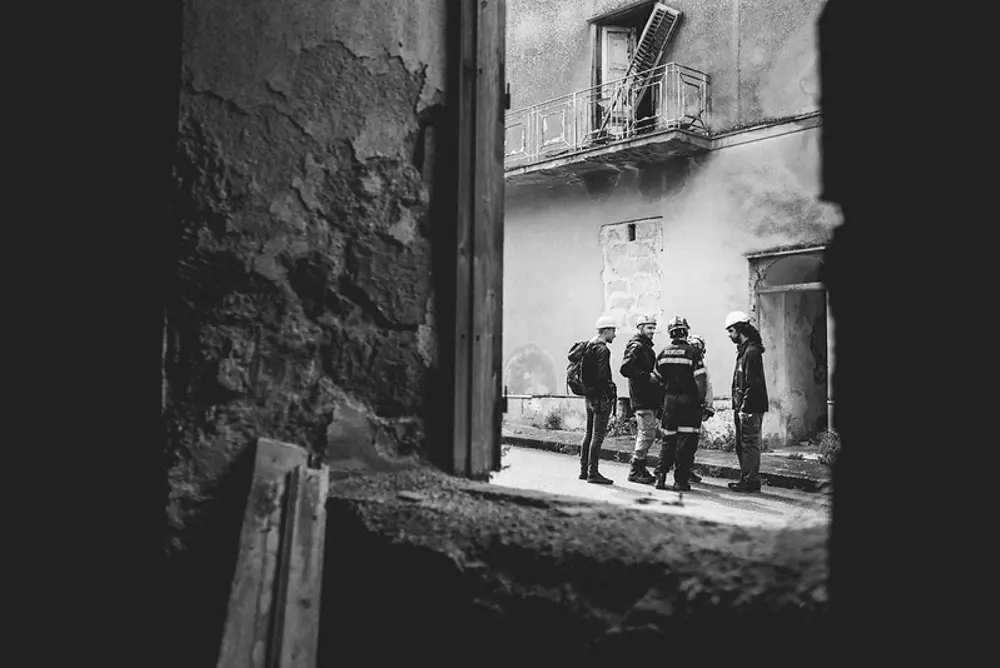 A group of people wearing helmets and protective gear stand together in a damaged street, seen through the crumbling window frame of a ruined building.