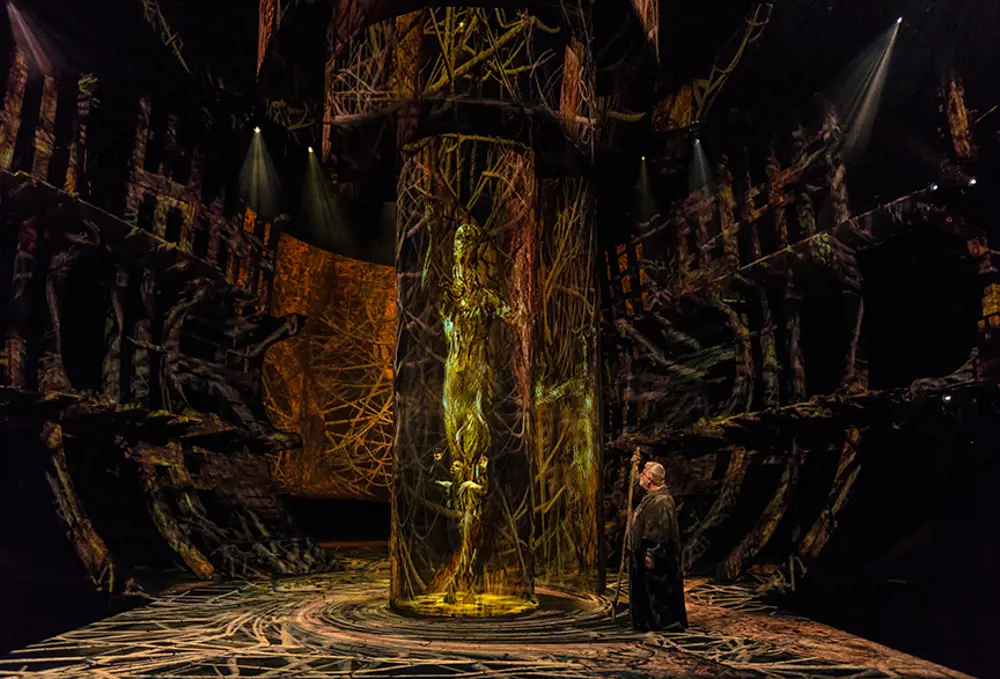 An actor with a staff, standing on stage in front of a gauze cylinder with special effects.