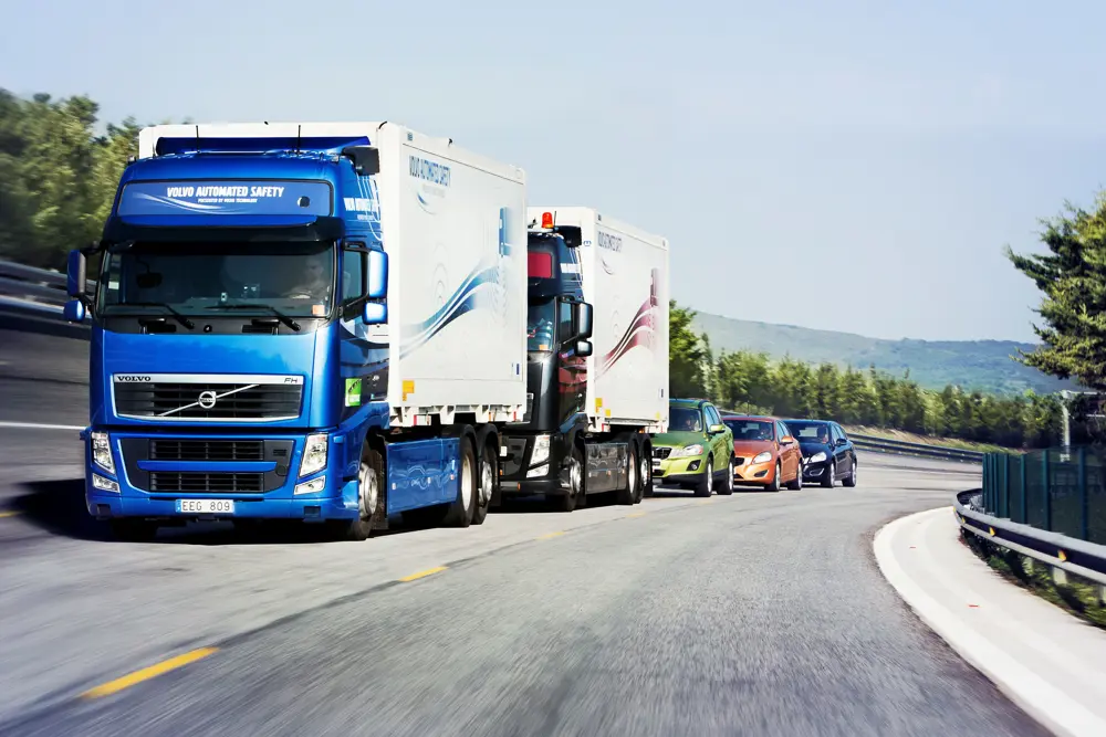 Two automated heavy goods vehicles following each other on a road, with three cars behind them.