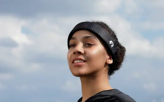 A Black woman with brown, tied up hair is wearing a black protective sports headband and a black t-shirt. Her face is tilted upwards and she is looking to the left of the camera