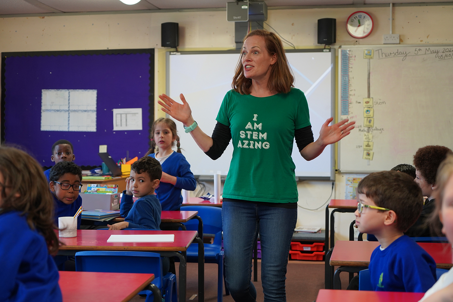 A woman in a green t-shirt and jeans is inside in a classroom talking to a mix gender group of students. The woman is gesturing as she speaks