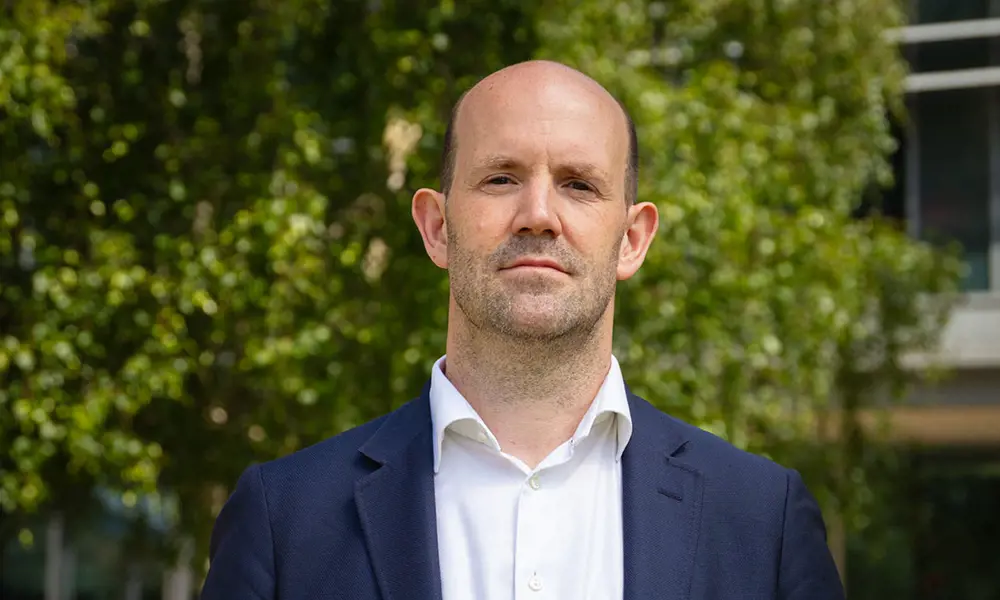 A man looks at the camera. He is wearing a white shirt and a navy blue blazer. He is standing outside with trees behind him