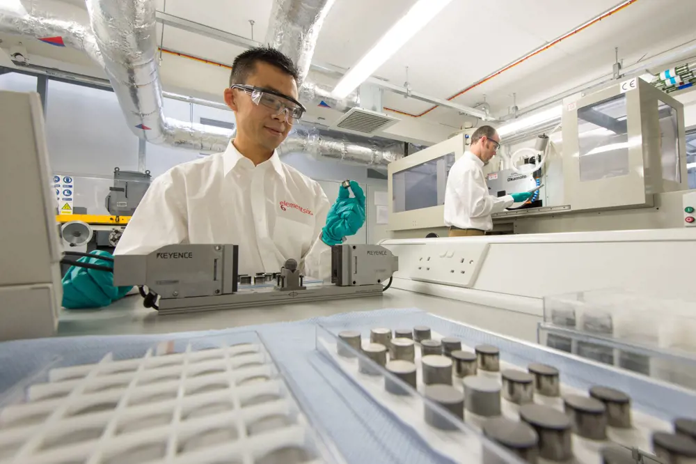 A person wearing gloves and glasses in a lab, holding a small cylinder.