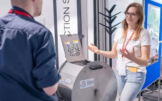 Person explaining a metal drum device during a demonstration, with another attendee listening and display boards in the background.