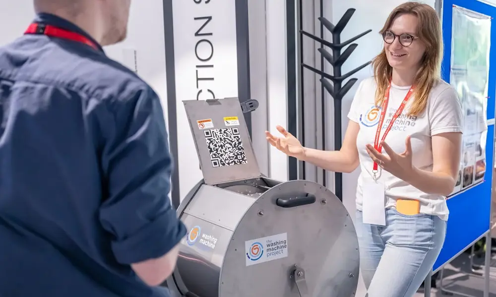 Person explaining a metal drum device during a demonstration, with another attendee listening and display boards in the background.
