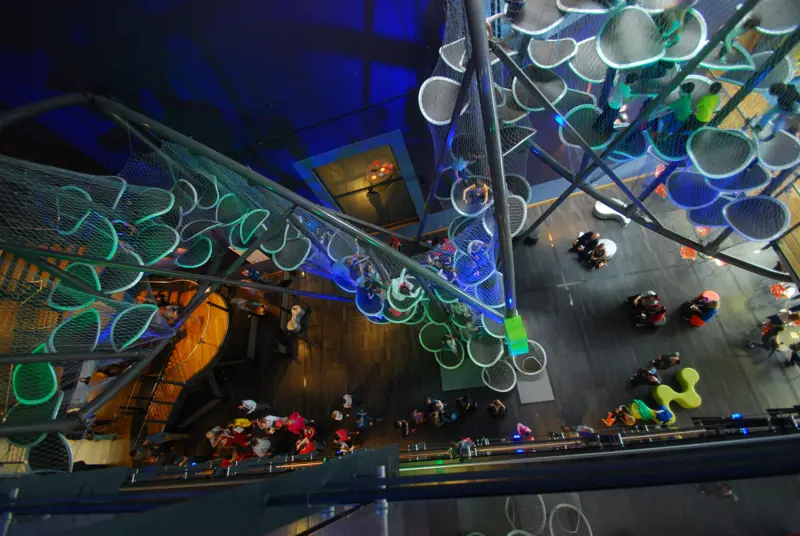 A birds eye view of a climbing frame in an atrium made of oval platforms contained in a mesh. People are walking around underneath