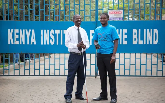 Brian Mwenda holding the sixth sense device and standing next to a visually impaired person smiling holding a white cane outside the gates of Kenya's Institute for the Blind.
