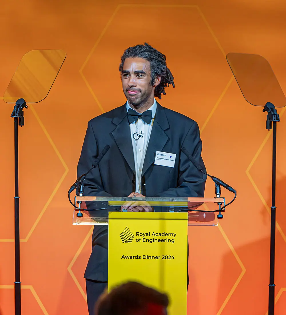 A person in formal evening wear stands at a clear lectern on a brightly lit stage at the Royal Academy of Engineering Awards Dinner 2024.