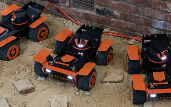 Three models of the orange and black Q-bot Spraybot robots, on a sandy floor next to a brick wall.
