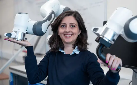 A woman with brown hair and a navy blue jumper posing for a photo, holding a robotic arm with each hand.
