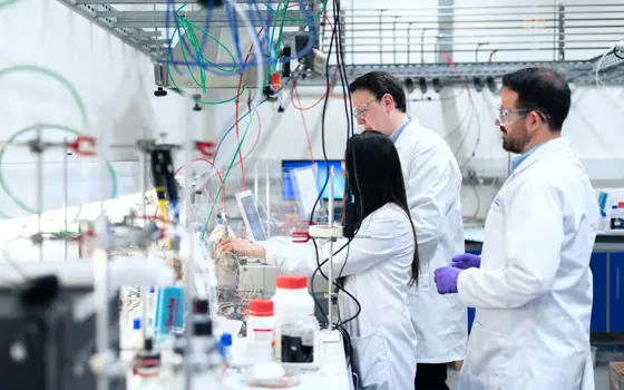 Three engineers in a lab, wearing lab coats and glasses, standing behind a desk full of science equipment, reagents and wires. 