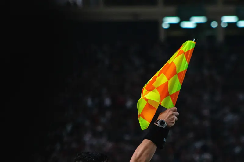 Assistant referee holding a fluorescent offside flag during a football match, with blurred stadium crowd behind.