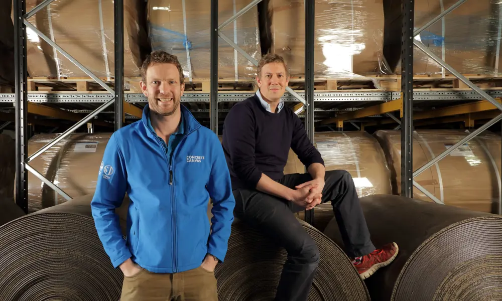 The two founders of Concrete Canvas, one sitting on a geotextile roll the size of a hay bale, the other standing in front wearing a blue zip through jacket.