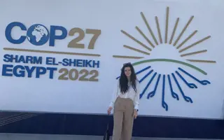 A woman standing in front of a sign for COP27 that says "Sharm El-Sheikh, Egypt 2022"