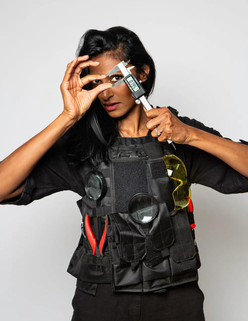 A woman engineering holding up a pair of calipers to her eye, she is wearing a black utility vest with other tools attached to it