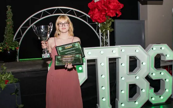A woman with blonde hair wearing glasses and a pink evening dress holds two certificates and a trophy.