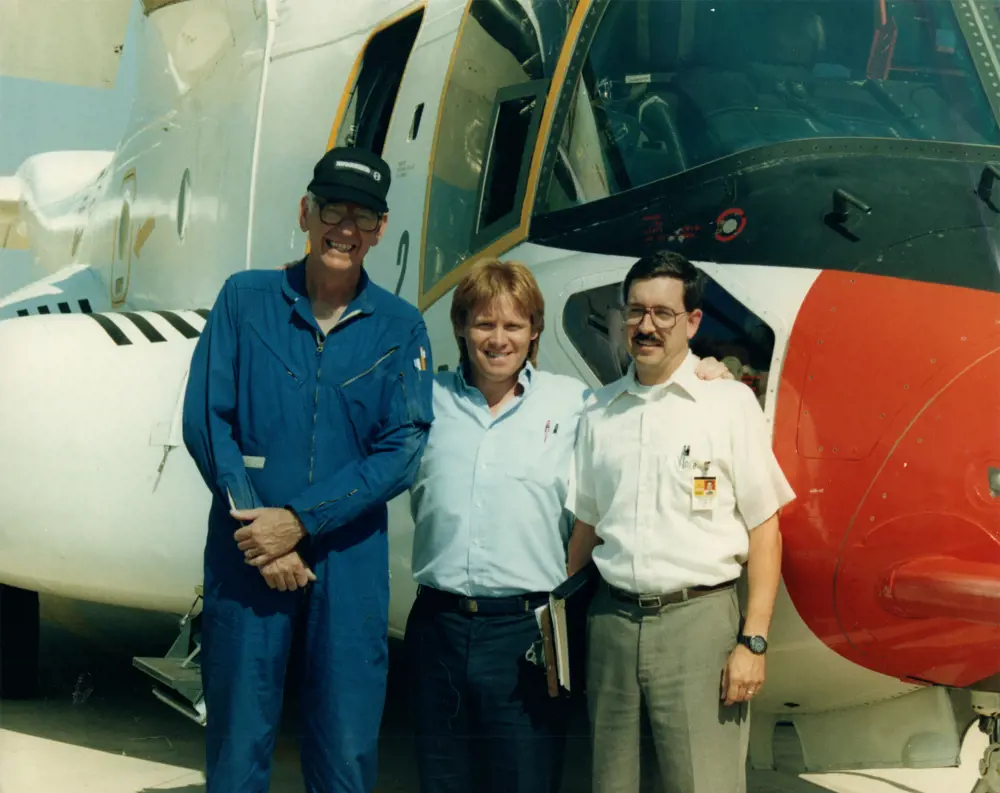 Three men standing in engineers' and pilots' uniforms, standing in front of a V22 aircraft.