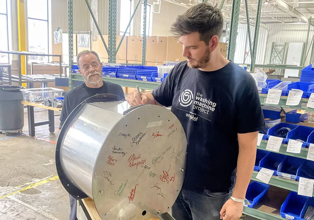 Two people in a workshop stand beside a large metal drum covered in handwritten signatures