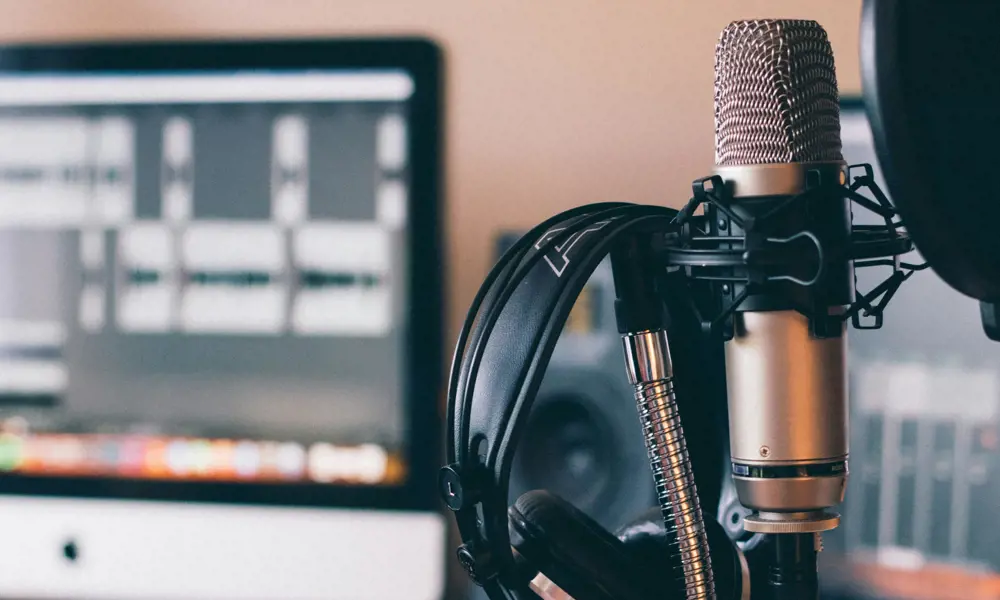 A microphone with headphones and a desk in the background with an iMac computer on it running an audio editing software.