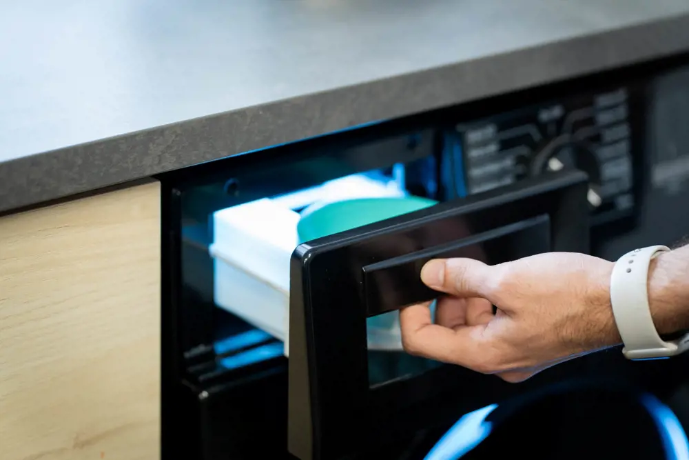 Image shows a washing machine drawer being pulled open by a person wearing a white watch - only their hand and wrist is seen