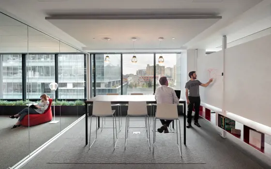A brightly lit office in San Francisco with one person pointing at a whiteboard and another sitting in a high chair at a table in the same room.