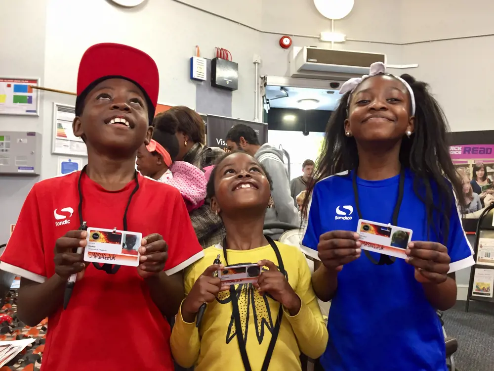 Three children smiling at the camera holding their future work ID cards, which are attached to a string around their necks.