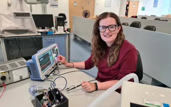 A photograph of Jean sitting at a desk adjusting the dial on an oscillator with one hand and holding a cable in the other.