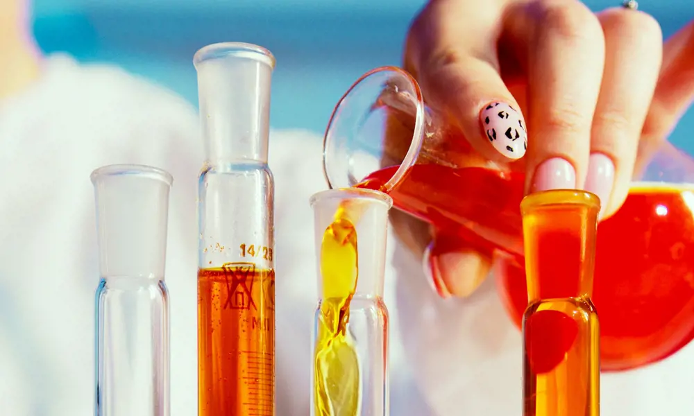 A manicured hand pouring chemicals into lab glassware.